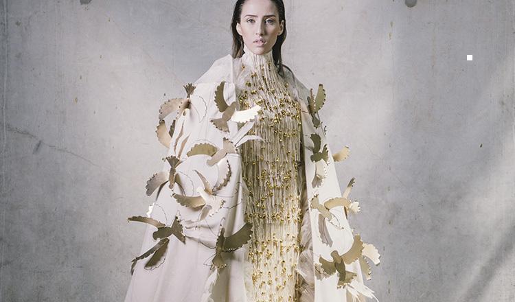 modelo luciendo un vestido con bordados dorados. Lleva una capa blanca con unas aves doradas en alto relieve que sobresalen