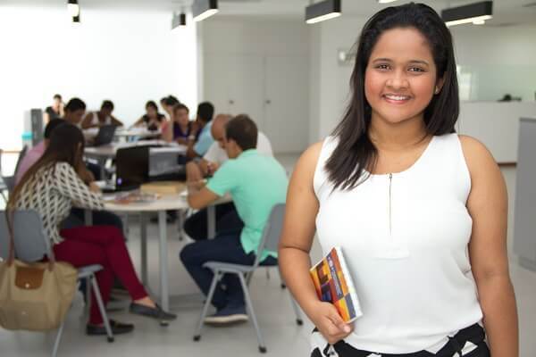 Jóven parada en primer plano con un cuaderno en la mano y al fondo se ve grupos de personas estudiando