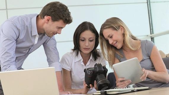 Tres estudiantes observan una cámara digital. Una de ellas sostiene una tableta. Al lado tiene un computador.